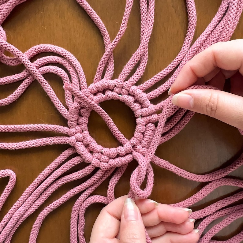 May include: A close-up of a pink macrame knotting project. The knotting is being done around a ring, and the ends of the cords are being held by hands.
