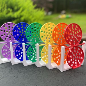 Può includere: Un set di giocattoli colorati a tema arcobaleno. Ogni giocattolo presenta un design circolare perforato in un colore diverso: viola, blu, verde, giallo, arancione e rosso. I giocattoli sono tenuti in un supporto rettangolare bianco.