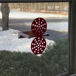 May include: A red, decorative, wall-mounted shelf with a unique design. The shelf features a rectangular base and a pair of circular, perforated elements. The shelf is mounted on a window, with a snowy outdoor scene visible in the background.
