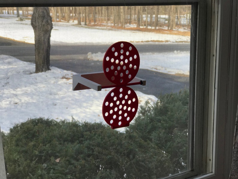 May include: A red, decorative, wall-mounted shelf with a unique design. The shelf features a rectangular base and a pair of circular, perforated elements. The shelf is mounted on a window, with a snowy outdoor scene visible in the background.