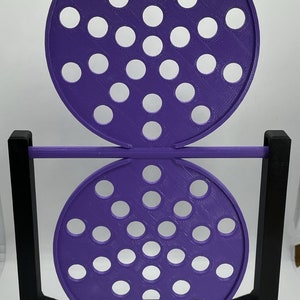 May include: A purple, 3D-printed object with two perforated circular discs connected by a horizontal rod. The discs have numerous white circular holes. The structure is supported by a black rectangular frame. The object is likely a decorative or functional item.