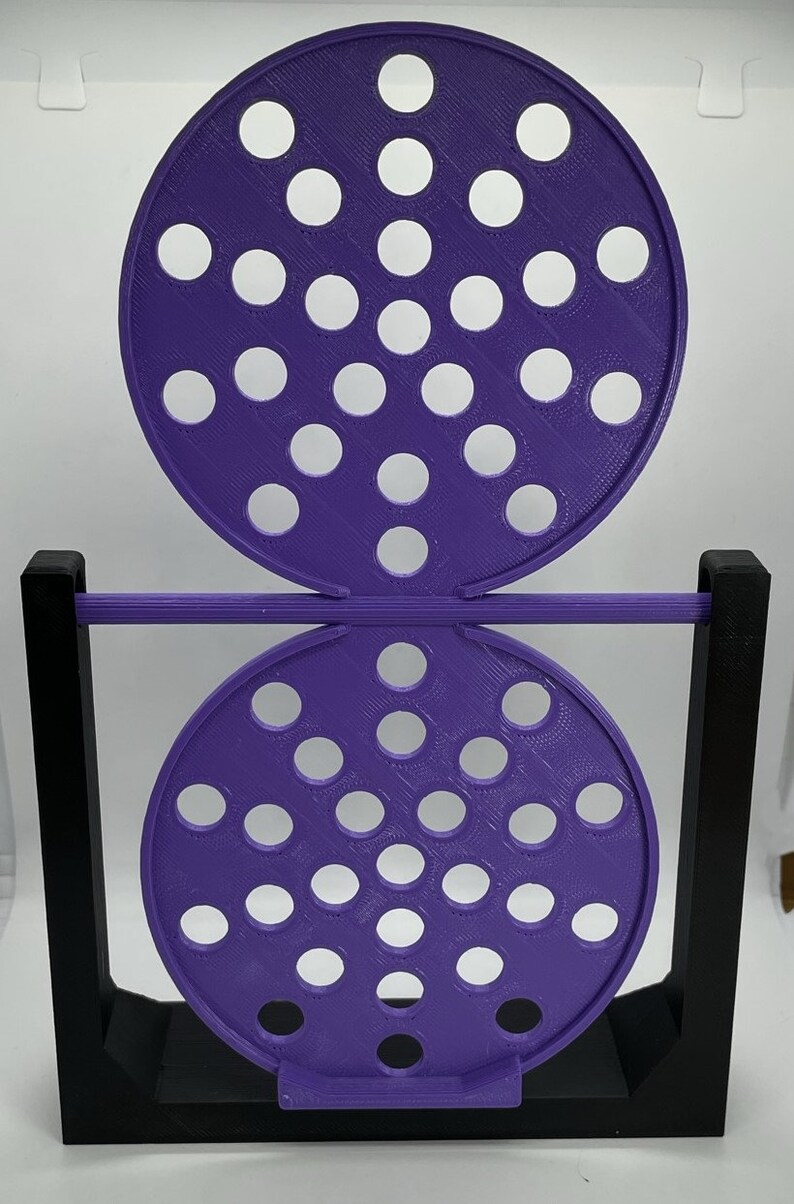 May include: A purple, 3D-printed object with two perforated circular discs connected by a horizontal rod. The discs have numerous white circular holes. The structure is supported by a black rectangular frame. The object is likely a decorative or functional item.