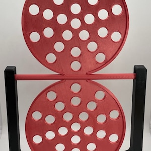 May include: A red, circular game with multiple holes, supported by a black frame. The game has two red circular pieces with evenly spaced holes, connected by a red bar. The black frame provides stability.