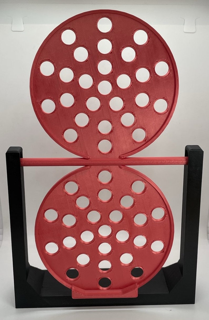 May include: A red, circular game with multiple holes, supported by a black frame. The game has two red circular pieces with evenly spaced holes, connected by a red bar. The black frame provides stability.