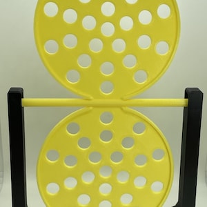 May include: A yellow and black 3D-printed game with two perforated circular pieces connected by a yellow bar. The game is supported by a black rectangular frame. The yellow pieces have multiple circular holes.