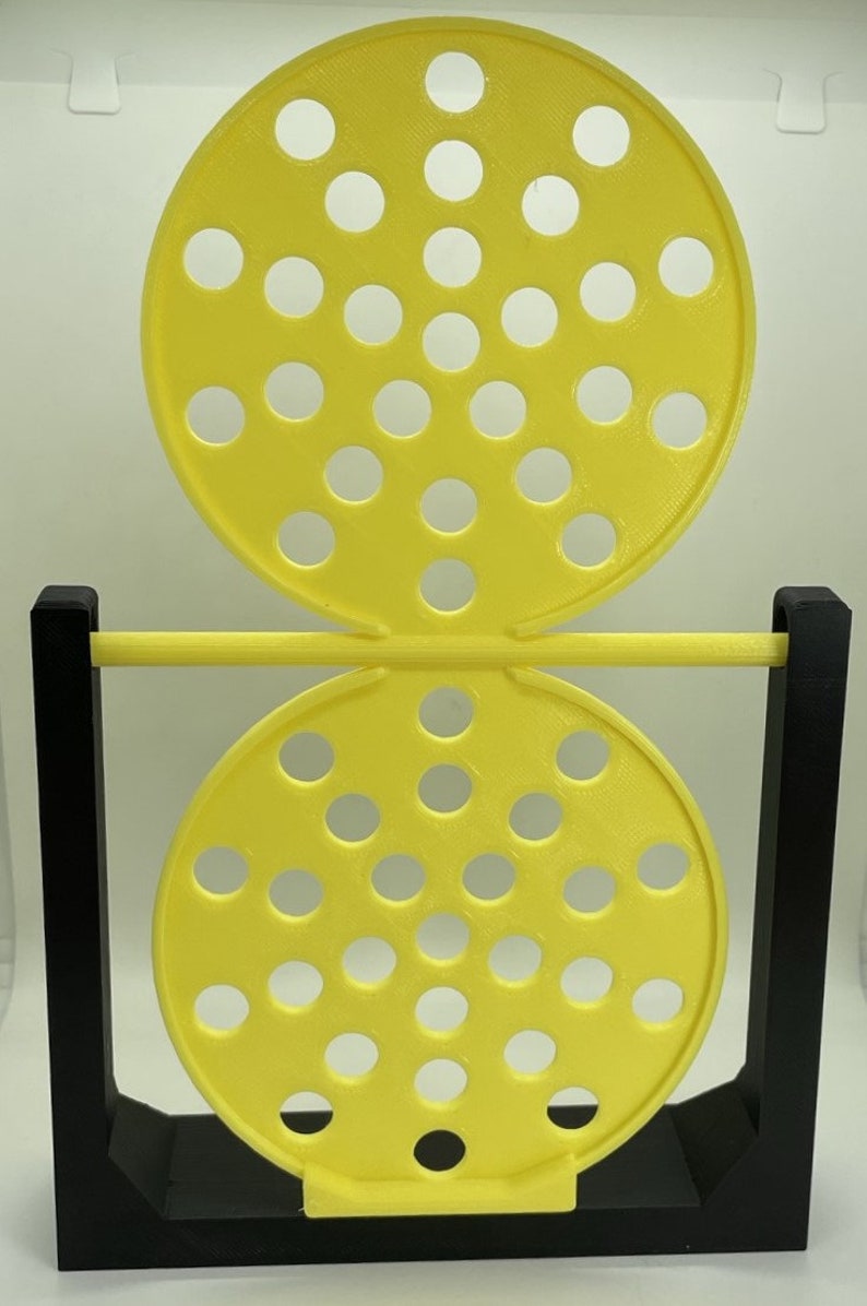 May include: A yellow and black 3D-printed game with two perforated circular pieces connected by a yellow bar. The game is supported by a black rectangular frame. The yellow pieces have multiple circular holes.