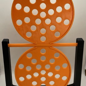 May include: An orange, circular, 3D-printed object with multiple holes, supported by a black frame. The object has two circular sections with evenly spaced holes, connected by a horizontal orange bar. The base is black.