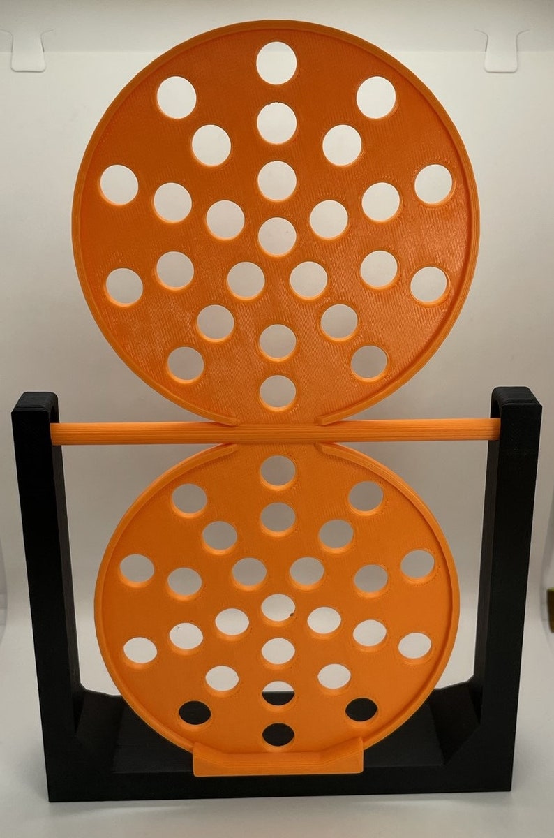 May include: An orange, circular, 3D-printed object with multiple holes, supported by a black frame. The object has two circular sections with evenly spaced holes, connected by a horizontal orange bar. The base is black.