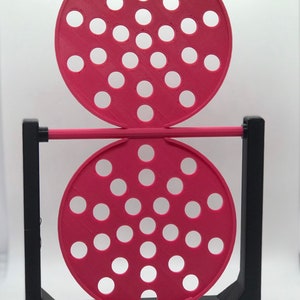 May include: A 3D-printed, geometric sculpture featuring two circular, perforated, magenta discs connected by a magenta rod. The discs are supported by a black, rectangular frame. The discs have multiple white circular holes.