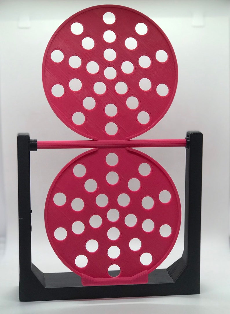 May include: A 3D-printed, geometric sculpture featuring two circular, perforated, magenta discs connected by a magenta rod. The discs are supported by a black, rectangular frame. The discs have multiple white circular holes.