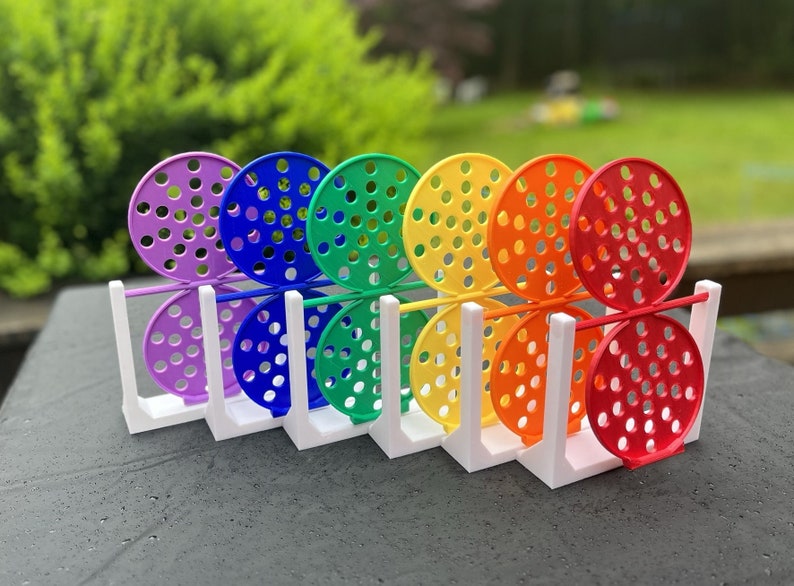 May include: A set of colorful, rainbow-themed toys. Each toy features a perforated, circular design in a different color: purple, blue, green, yellow, orange, and red. The toys are held in a white, rectangular stand, creating a vibrant display.