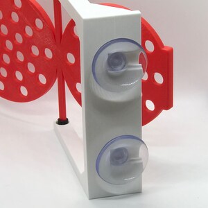May include: A white and red 3D-printed butterfly-shaped object with suction cups. The butterfly has a series of circular cutouts. The white section has two clear suction cups. The object is designed to be mounted on a smooth surface.