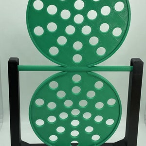 May include: A green, circular, perforated object is suspended between two black rectangular supports. The object is composed of two connected discs, each with numerous evenly spaced holes. The design is a modern, geometric sculpture.