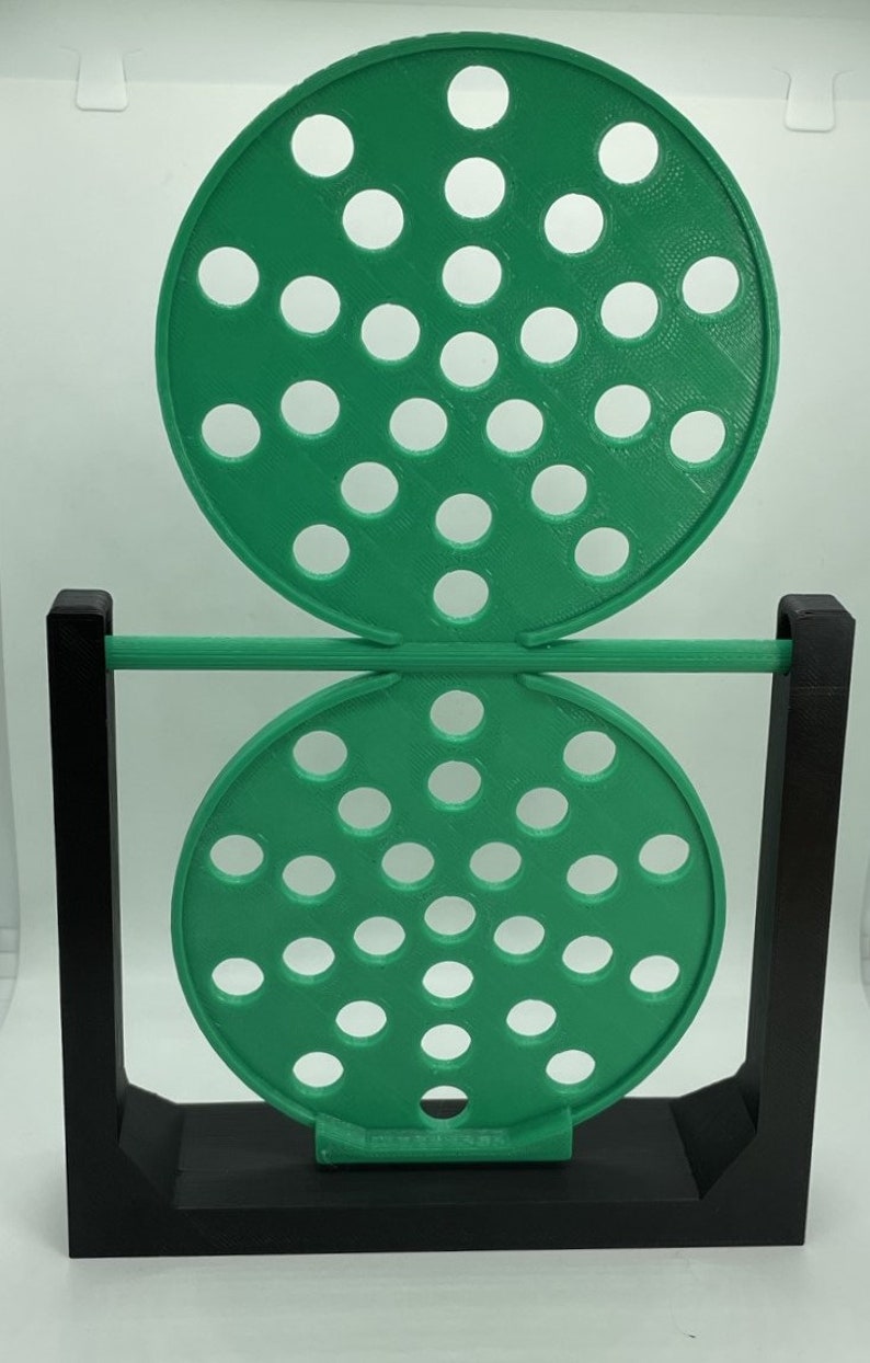 May include: A green, circular, perforated object is suspended between two black rectangular supports. The object is composed of two connected discs, each with numerous evenly spaced holes. The design is a modern, geometric sculpture.