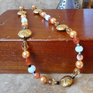 May include: A long necklace with orange, peach, white, and gold beads. The necklace has a decorative gold charm with a floral design.