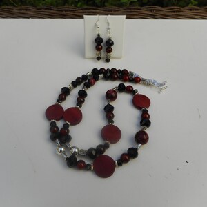 Puede incluir: Un collar de dos hilos con cuentas de madera roja, cuentas negras y detalles plateados. Pendientes a juego con cuentas negras y rojas.