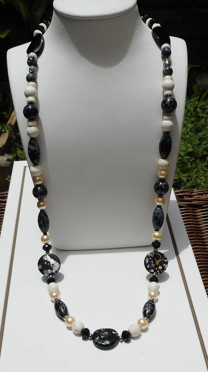 May include: A long black, white, and gold beaded necklace with oval and round beads. The necklace is made of various materials, including stone and metal.