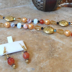 May include: A long necklace with gold and orange beads, and matching earrings with orange beads. The necklace has a gold clasp and a decorative gold pendant.