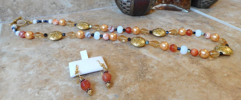 May include: A long necklace with gold and orange beads, and matching earrings with orange beads. The necklace has a gold clasp and a decorative gold pendant.