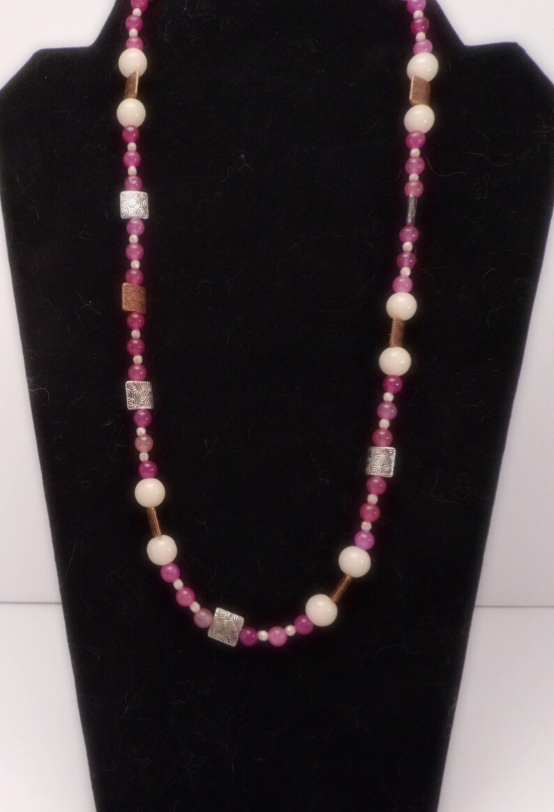 Pode incluir: Um colar de contas rosa e branco com detalhes prateados e de cobre. O colar &eacute; feito com contas redondas e quadradas.