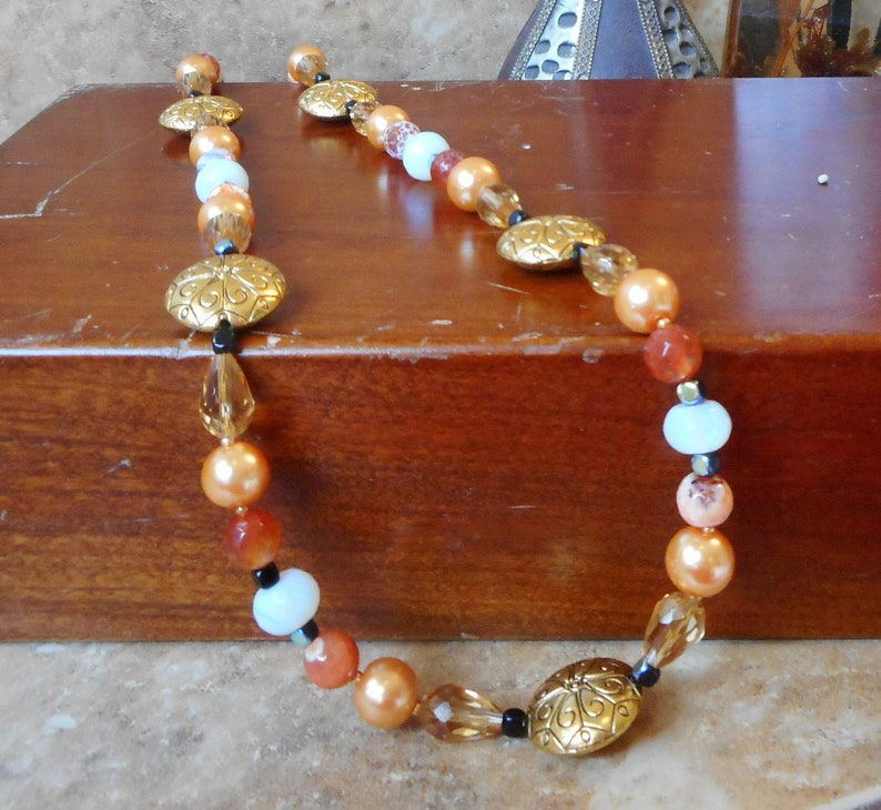 May include: A long necklace with orange, peach, white, and gold beads. The necklace has a decorative gold charm with a floral design.