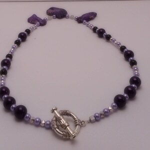 May include: A purple and silver beaded necklace with a silver toggle clasp. The necklace features a variety of purple beads, including round and oval shapes.
