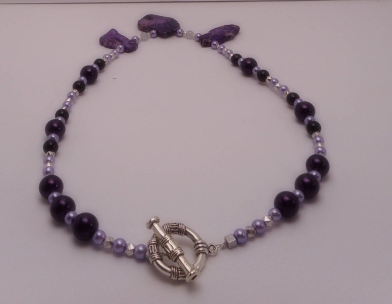 May include: A purple and silver beaded necklace with a silver toggle clasp. The necklace features a variety of purple beads, including round and oval shapes.