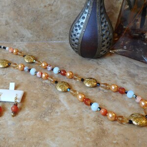 May include: A long necklace with gold and brown beads, featuring a repeating pattern of gold oval charms. The necklace is paired with matching earrings with a single brown bead.