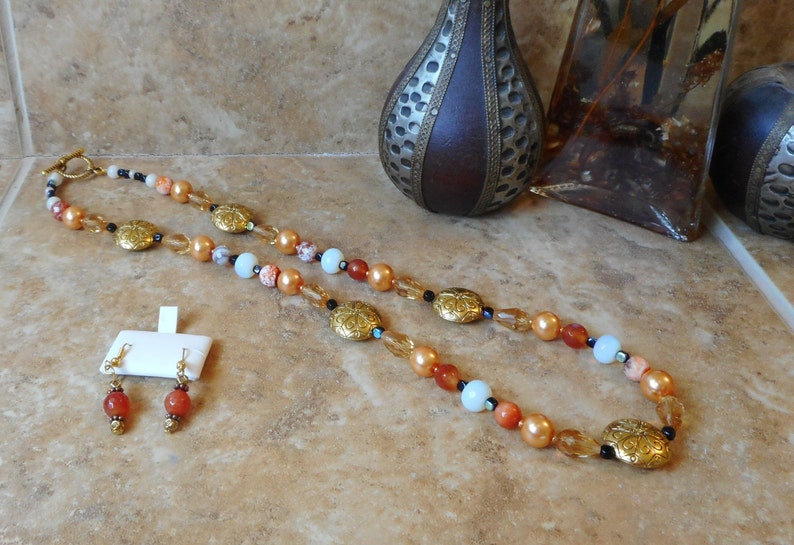 May include: A long necklace with gold and brown beads, featuring a repeating pattern of gold oval charms. The necklace is paired with matching earrings with a single brown bead.