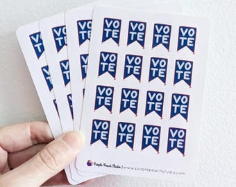 Op de afbeelding: Een hand die een stapel witte stickers vasthoudt. Elke sticker heeft een blauw bannerontwerp met het woord "VOTE" in het wit. De stickers zijn van Purple Peach Studio.