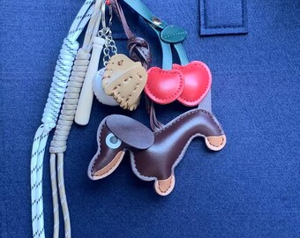 May include: A blue felt tote bag with a variety of charms attached to the handle. The charms include a baseball, a dachshund, cherries, and a white and beige rope.