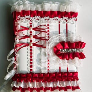 May include: A white and red decorative item with layers of ruffles, ribbons, and lace. The design features red satin ribbons, white lace trim, and faux pearls. A matching red and white accessory with a rhinestone band is also included.