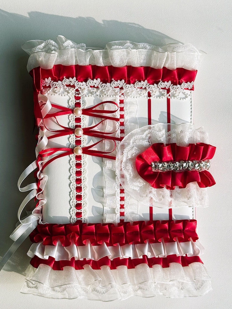May include: A white and red decorative item with layers of ruffles, ribbons, and lace. The design features red satin ribbons, white lace trim, and faux pearls. A matching red and white accessory with a rhinestone band is also included.