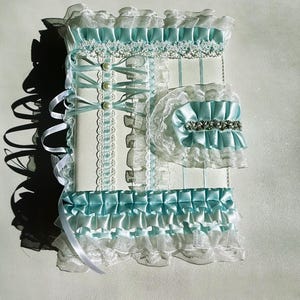 May include: A decorative, white and light blue journal with lace and ribbon accents. The journal features a matching garter with a silver embellishment. The design includes pearls and ruffled edges.