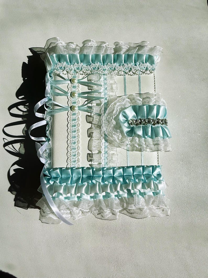 May include: A decorative, white and light blue journal with lace and ribbon accents. The journal features a matching garter with a silver embellishment. The design includes pearls and ruffled edges.