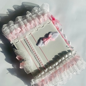 May include: A white, rectangular item with pink and white frills and ribbons. The item has a decorative lace trim and a small bow. The overall design is delicate and feminine.