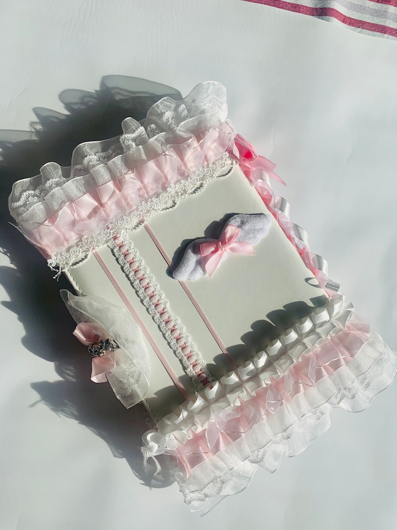 May include: A white, rectangular item with pink and white frills and ribbons. The item has a decorative lace trim and a small bow. The overall design is delicate and feminine.