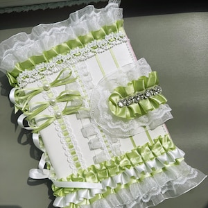 May include: A white and green decorative journal with lace and ribbon accents. The journal features a layered design with green satin ribbon, white lace trim, and pearl embellishments. A matching accessory with a rhinestone accent is also included.