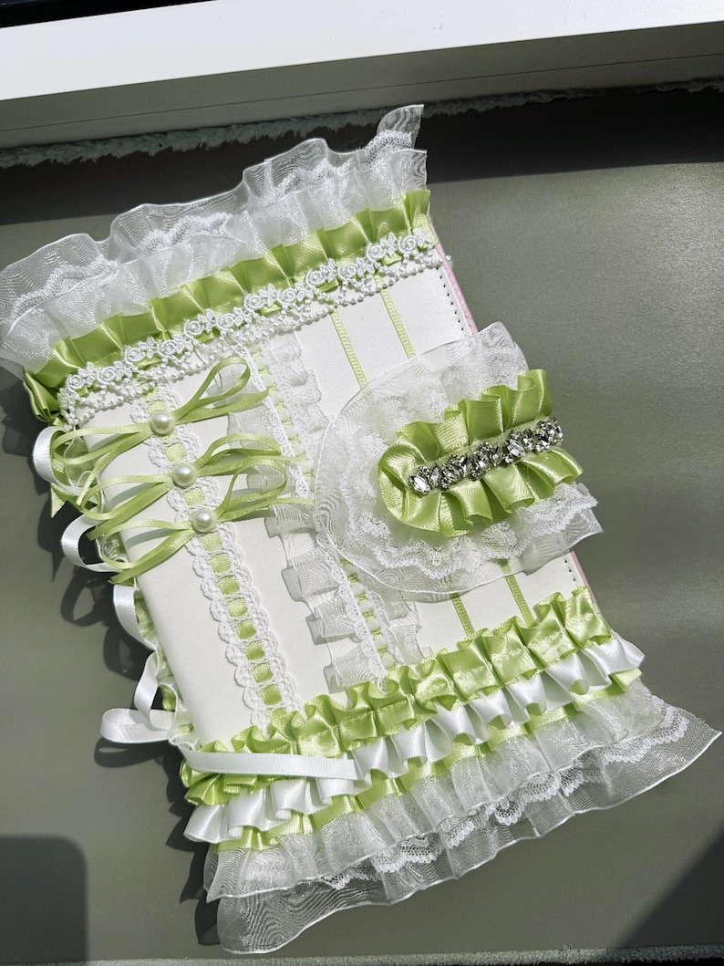 May include: A white and green decorative journal with lace and ribbon accents. The journal features a layered design with green satin ribbon, white lace trim, and pearl embellishments. A matching accessory with a rhinestone accent is also included.