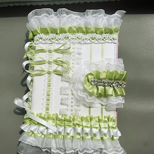 May include: A decorative, handmade journal with a white cover, adorned with layers of white lace, light green satin ribbons, and pearl accents. The journal features a ruffled trim and a matching embellishment with rhinestones.