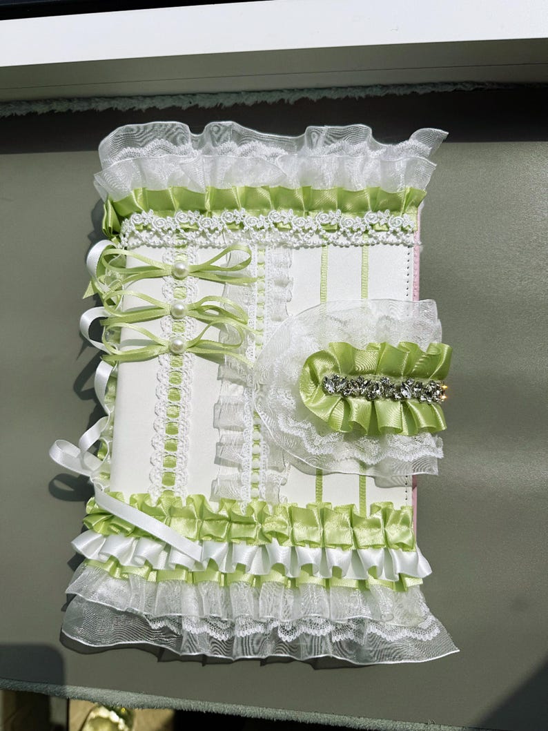May include: A decorative, handmade journal with a white cover, adorned with layers of white lace, light green satin ribbons, and pearl accents. The journal features a ruffled trim and a matching embellishment with rhinestones.