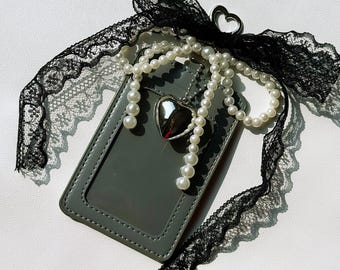 May include: A gray cardholder with a clear window, adorned with a silver heart charm and a strand of faux pearls. A black lace ribbon and a heart-shaped metal loop complete the design, creating a romantic aesthetic.