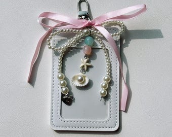May include: White ID card holder with a pearl and seashell charm, a pink ribbon, and a silver clasp. The charm includes a small seashell, a starfish, and beads in shades of pink and blue. The card holder is made of a smooth, white material.