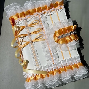 May include: A white and yellow decorative journal with a layered design. The journal features white lace, yellow satin ribbons, and pearl accents. The edges are trimmed with ruffled fabric, and the cover has a bow closure.