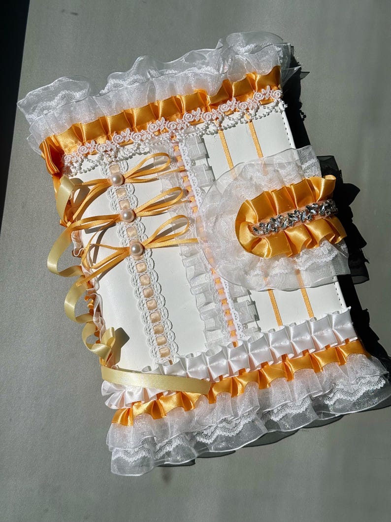 May include: A white and yellow decorative journal with a layered design. The journal features white lace, yellow satin ribbons, and pearl accents. The edges are trimmed with ruffled fabric, and the cover has a bow closure.