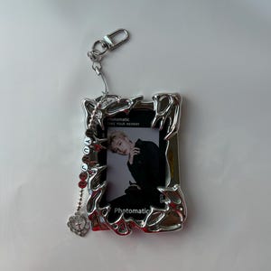 May include: Silver photo frame keychain with a silver chain and a small silver heart charm. The frame has a photo of a person inside. The text "Photomatic Take Your Memory" is printed on the frame.