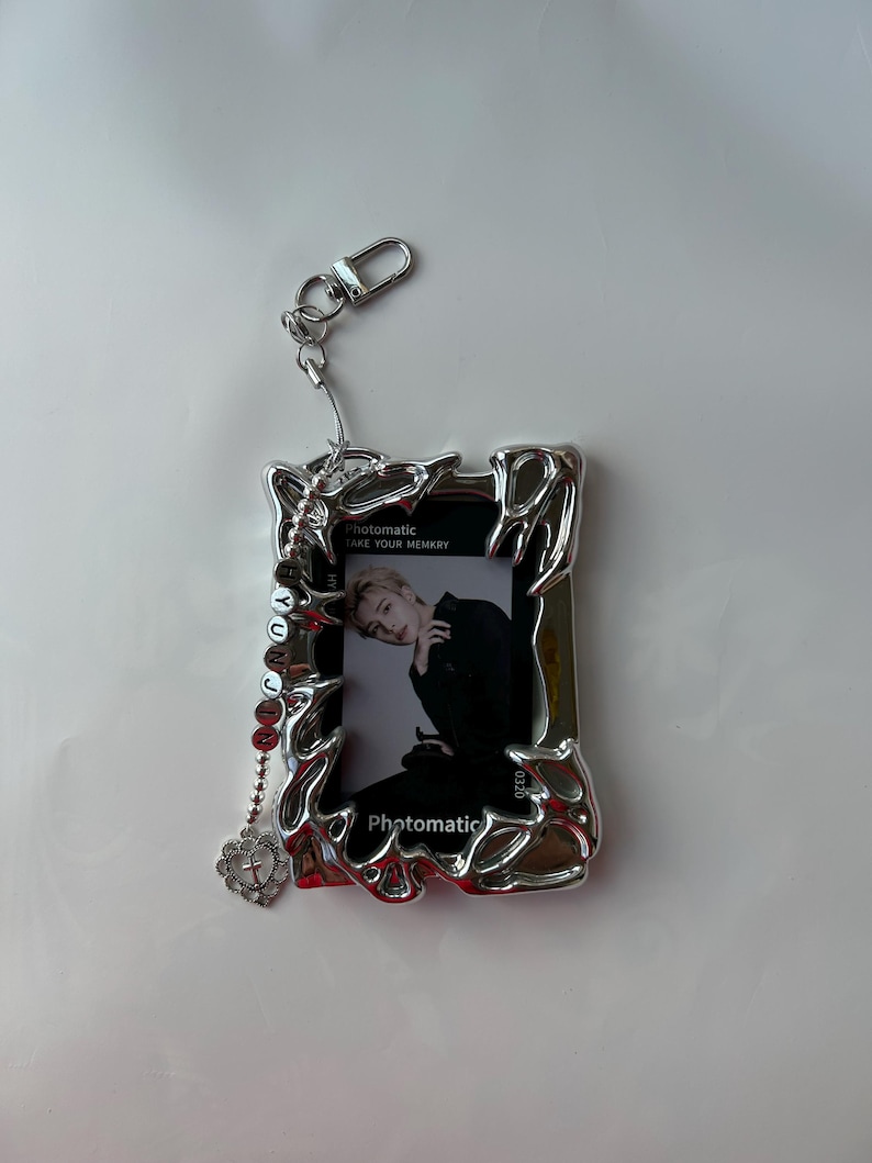 May include: Silver photo frame keychain with a silver chain and a small silver heart charm. The frame has a photo of a person inside. The text "Photomatic Take Your Memory" is printed on the frame.