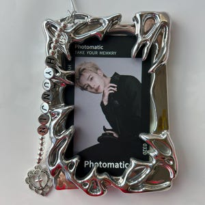 May include: A silver photo frame with a jagged, organic design. The frame has a photo inside of a young man with blonde hair. The photo is from the Photomatic app and says "Take Your Memkry". A silver chain with a charm that says "HYUNJIN" hangs from the top of the frame.