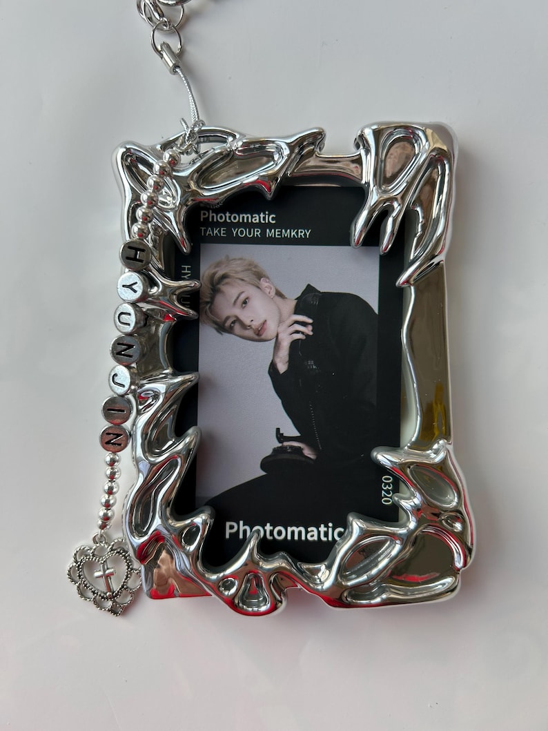 May include: A silver photo frame with a jagged, organic design. The frame has a photo inside of a young man with blonde hair. The photo is from the Photomatic app and says "Take Your Memkry". A silver chain with a charm that says "HYUNJIN" hangs from the top of the frame.