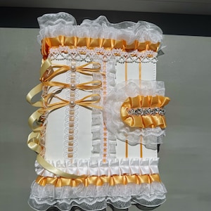 May include: A white and yellow decorative journal with lace and ribbon accents. The journal features a ruffled trim, satin ribbons, and pearl embellishments. The design includes a matching accessory with a jeweled band.
